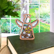 The Forged Flare® White Plumeria Ornament | 3.5" (H) Mother's Angels® angel figurine sits on a closed green book near a window, serving as a charming Hawaiian Christmas decoration overlooking a lush garden.