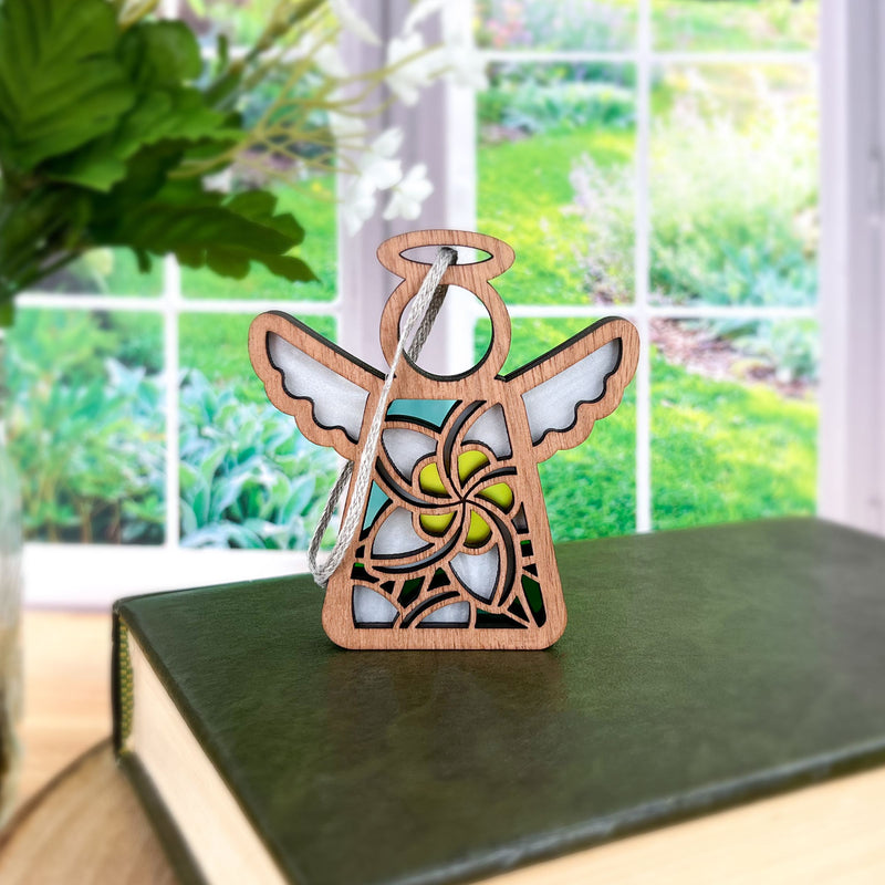 The Forged Flare® White Plumeria Ornament | 3.5" (H) Mother's Angels® angel figurine sits on a closed green book near a window, serving as a charming Hawaiian Christmas decoration overlooking a lush garden.