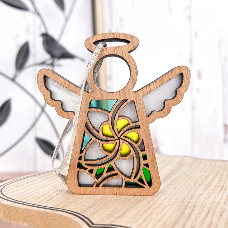 The Forged Flare® White Plumeria Ornament is a 3.5" Mother's Angels® figurine with cutout wings and stained glass-style yellow and green accents, standing on light wood with a string for hanging—a unique Hawaiian holiday gift.