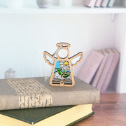 The Forged Flare® Book Lovers Ornament, a 3.5" tall Mother's Angels® figurine with a stained-glass floral design, sits atop two books—making it a charming book club gift. Softly blurred shelves and a row of books complete the background.