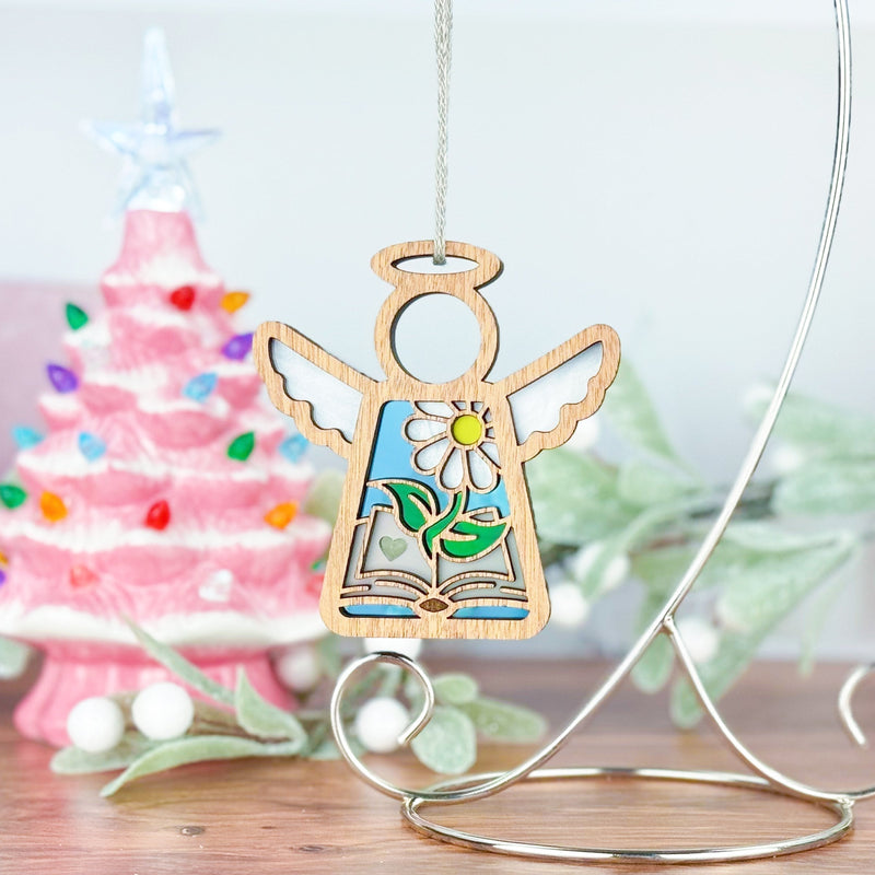 The Forged Flare® Book Lovers Ornament—a 3.5" Mother's Angels® angel figurine adorned with daisies and leaves—hangs on a silver stand, with a pink Christmas tree and festive greenery creating an ideal gift scene for book lovers.