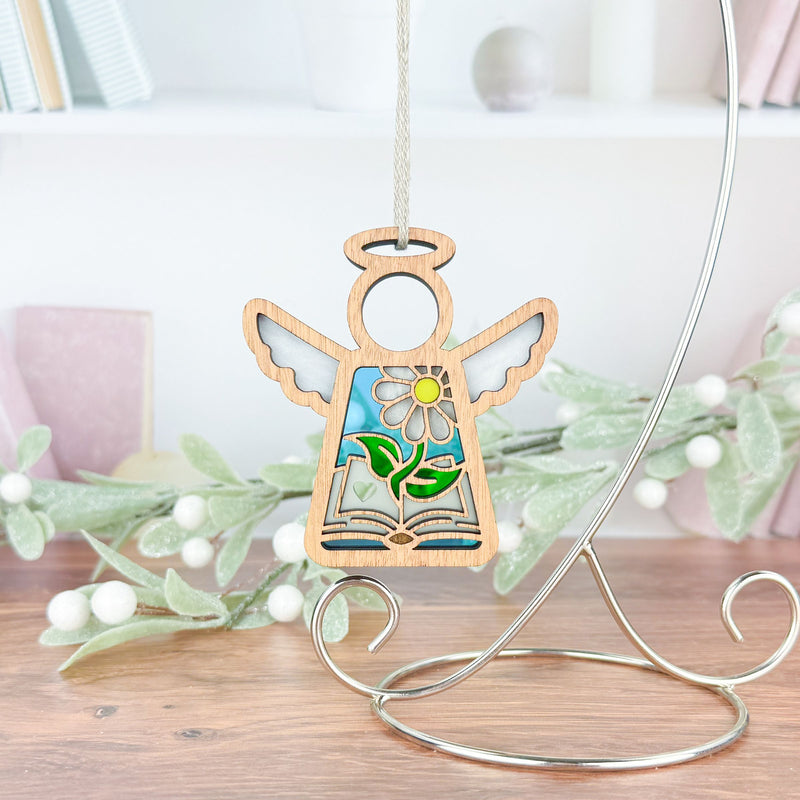 The Forged Flare® Book Lovers Ornament—a 3.5" Mother's Angels® wooden angel figurine with stained glass wings and a sunflower design—hangs from a silver stand, making a charming book club gift. White faux berries and leaves accent the background.