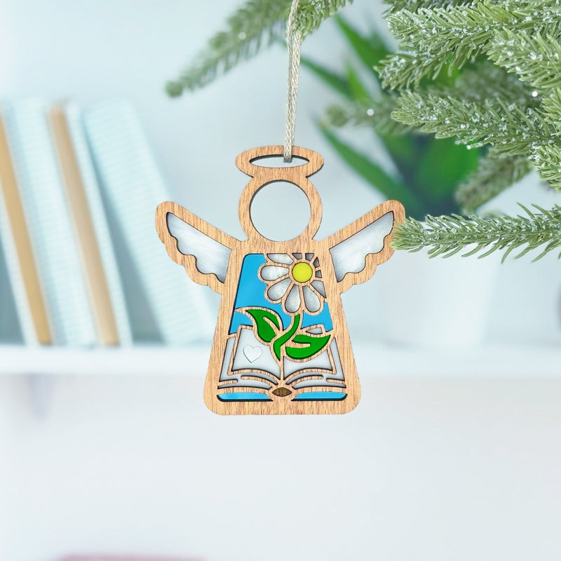 The Forged Flare® Book Lovers Ornament—a 3.5" Mother's Angels® wooden angel with a flower and leaf motif—hangs on a green Christmas tree branch, making a perfect book club gift. Blurred books and a plant appear in the background.