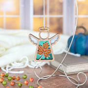 The Forged Flare® Sugar Maple Leaf for Charity Ornament | 3.5" (H) Angel Figurine by Mother's Angels® features white wings and a golden autumn leaf, suspended from a silver stand with warm Appalachian Mountains light shining through a window behind it.
