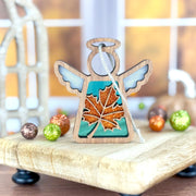 A Forged Flare® Mother's Angels® 3.5" Sugar Maple Leaf for Charity Angel Ornament stands on a tray with colorful balls, blue wings gleaming, as an autumn Appalachian scene appears through a blurred window.
