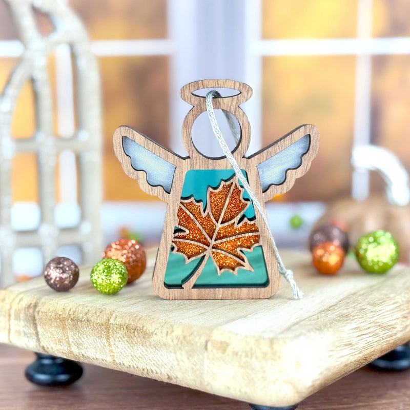 A Forged Flare® Mother's Angels® 3.5" Sugar Maple Leaf for Charity Angel Ornament stands on a tray with colorful balls, blue wings gleaming, as an autumn Appalachian scene appears through a blurred window.