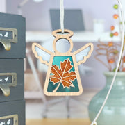 The Forged Flare® Sugar Maple Leaf for Charity Ornament, a 3.5" Mother's Angels® figurine with white wings and a teal dress glittered with an orange maple leaf, evokes the Appalachian spirit against a soft-focus office backdrop.