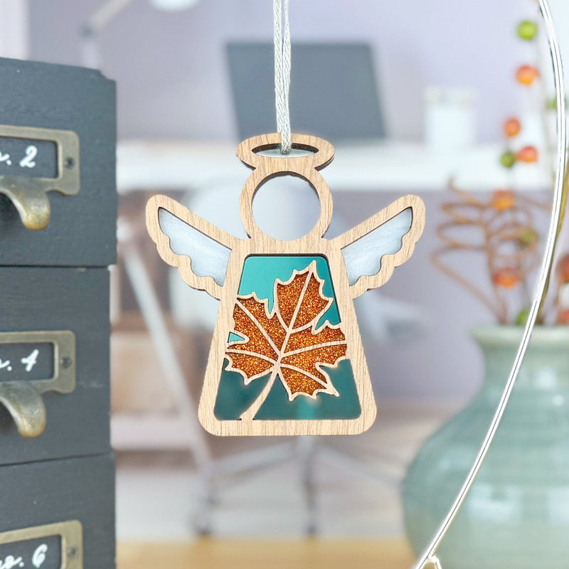 The Forged Flare® Sugar Maple Leaf for Charity Ornament, a 3.5" Mother's Angels® figurine with white wings and a teal dress glittered with an orange maple leaf, evokes the Appalachian spirit against a soft-focus office backdrop.