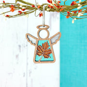 The Forged Flare® Sugar Maple Leaf for Charity Ornament, a 3.5" Mother's Angels® angel figurine, hangs gracefully from a decorative branch with orange and white berries, reflecting Appalachian inspiration and warm Samaritan’s Purse support.