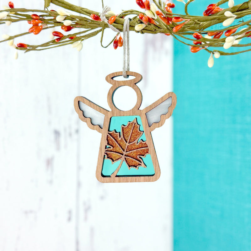 The Forged Flare® Sugar Maple Leaf for Charity Ornament, a 3.5" Mother's Angels® angel figurine, hangs gracefully from a decorative branch with orange and white berries, reflecting Appalachian inspiration and warm Samaritan’s Purse support.