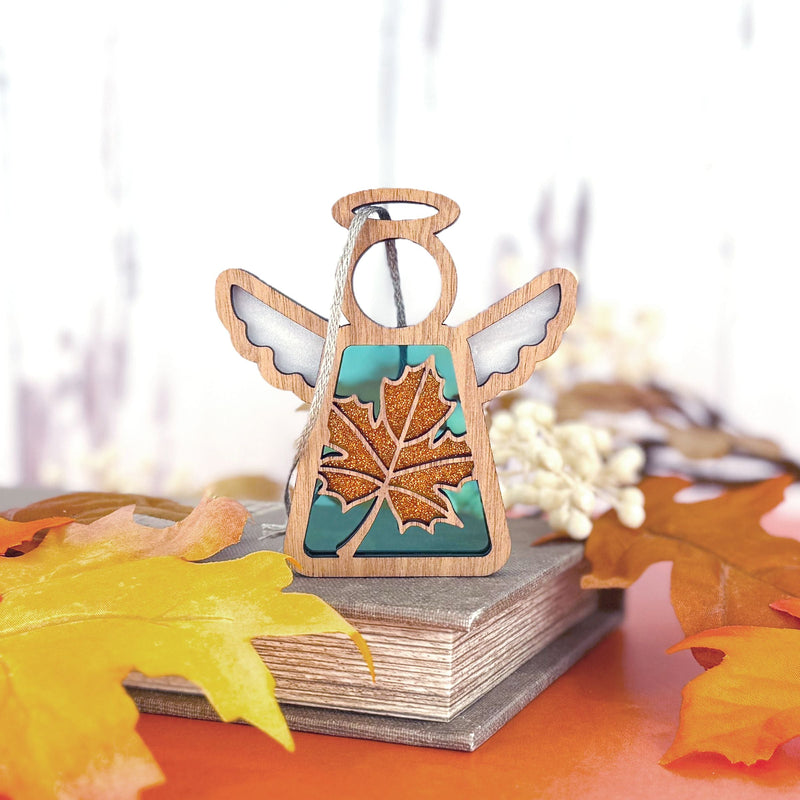 A Forged Flare® Sugar Maple Leaf for Charity Ornament | 3.5" (H) Angel Figurine from the Mother's Angels® collection stands on a closed book amid autumn leaves, with a cozy, Appalachian-inspired atmosphere in the softly blurred background.