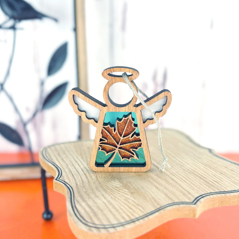 The Forged Flare® Sugar Maple Leaf for Charity Ornament | 3.5" (H) Mother's Angels® angel figurine features white wings, a halo, and a brown maple leaf on a wooden stand with an orange background—honoring the spirit of the Appalachians.