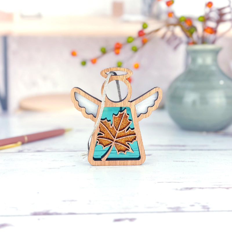 The Forged Flare® Sugar Maple Leaf for Charity Ornament, a 3.5" Mother's Angels® angel figurine with a gold maple leaf and turquoise accents inspired by the Appalachian Mountains, stands on a white surface with blurred orange and green berries behind.