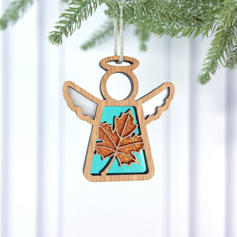 The Forged Flare® Sugar Maple Leaf for Charity Ornament | 3.5" (H) Angel Figurine by Mother's Angels® features a glittery orange maple leaf and turquoise center, hanging from a pine branch against a white background, inspired by the Appalachians.