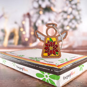 A PREORDERS CLOSED! Poinsettia Ornament | 3.5" (H) Angel Figurine by Forged Flare® from the Mother's Angels® collection stands on a gift box, with softly blurred Christmas lights, tree decor, and reindeer shapes in the background.