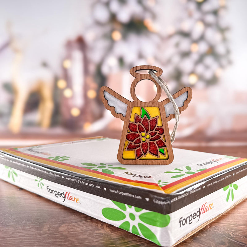 A PREORDERS CLOSED! Poinsettia Ornament | 3.5" (H) Angel Figurine by Forged Flare® from the Mother's Angels® collection stands on a gift box, with softly blurred Christmas lights, tree decor, and reindeer shapes in the background.