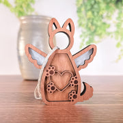 The Forged Flare® Forever By My Side Dog Angel Ornament features a winged dog with a heart and paw prints, stands on wood, and comes with a hanging string. A blurred vase with green plants is visible in the background.