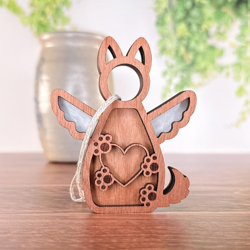 The Forged Flare® Forever By My Side Dog Angel Ornament features a winged dog with a heart and paw prints, stands on wood, and comes with a hanging string. A blurred vase with green plants is visible in the background.