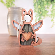The Forever By My Side Dog Angel Ornament by Forged Flare® is a winged dog-shaped keepsake with heart and paw cutouts, displayed on wood—a thoughtful gift for dog lovers, featuring a string, with a vase and greenery blurred in the background.