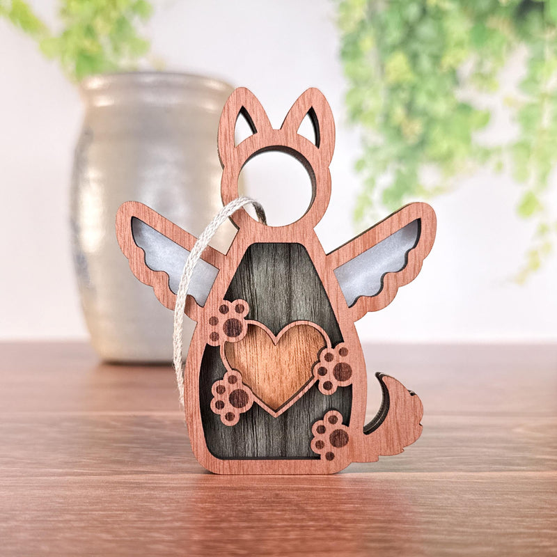 The Forever By My Side Dog Angel Ornament by Forged Flare® is a winged dog-shaped keepsake with heart and paw cutouts, displayed on wood—a thoughtful gift for dog lovers, featuring a string, with a vase and greenery blurred in the background.