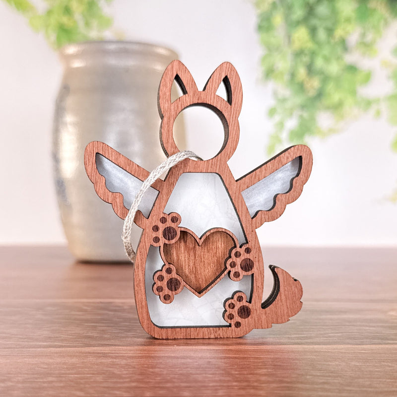 The Forged Flare® Forever By My Side Dog Angel Ornament features a winged dog with a heart and paw prints, standing on wood. A silver vase and blurred green foliage appear in the background.
