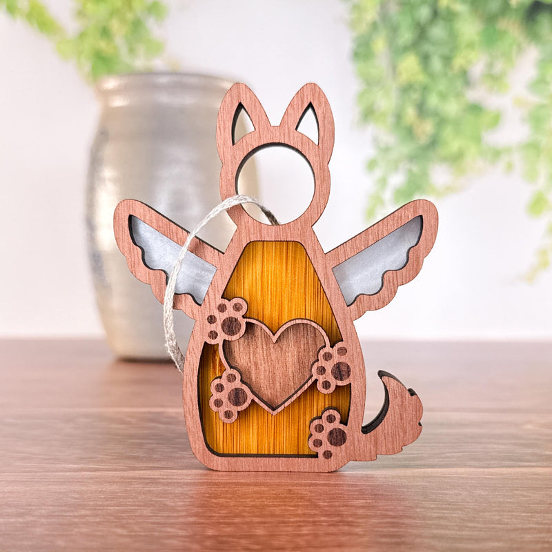 The Forever By My Side Dog Angel Ornament by Forged Flare® is a wooden winged dog with heart and paw prints, finished with a silver hanging string—a perfect dog lover gift. A blurred vase and green plants are in the background.