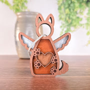 The Forged Flare® Forever By My Side Dog Angel Ornament—a wooden decoration with a winged dog, heart, and paw prints cutout—leans by a vase on wood; perfect as a dog lover gift or memorial with blurred green plants in the background.