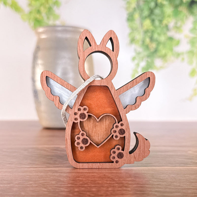 The Forged Flare® Forever By My Side Dog Angel Ornament—a wooden decoration with a winged dog, heart, and paw prints cutout—leans by a vase on wood; perfect as a dog lover gift or memorial with blurred green plants in the background.