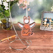 The Forged Flare® Forever By My Side Dog Angel Ornament, with wings and a heart on its chest, hangs from a metal stand on a wooden table—an ideal dog lover gift. A vase of flowers and a framed photo of three people sit in the background.