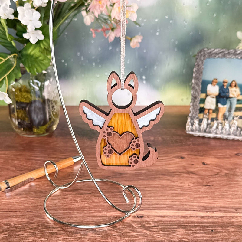 The Forged Flare® Forever By My Side Dog Angel Ornament, featuring a winged dog with a heart on its chest, hangs from a metal stand on a wooden table—an ideal gift for dog lovers. Flowers and a family photo decorate the background.