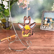 The Forged Flare® Forever By My Side Dog Angel Ornament hangs from a metal stand on a wooden table, making a heartfelt dog memorial and thoughtful dog lover gift. Nearby are flowers in a vase, a pen, and a beach photo with three people.