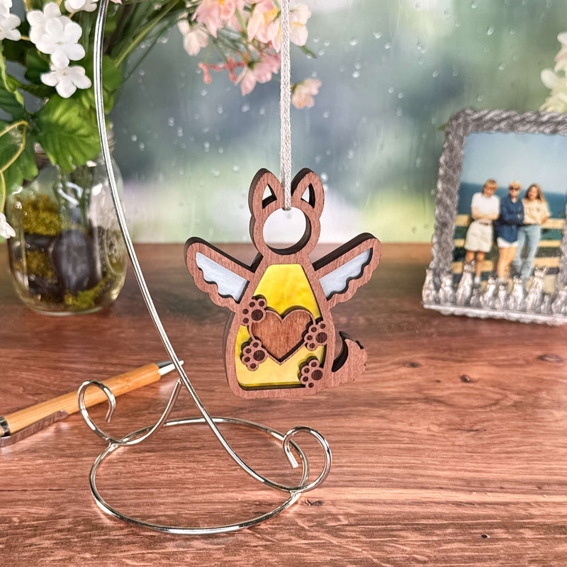 The Forged Flare® Forever By My Side Dog Angel Ornament hangs from a metal stand on a wooden table, making a heartfelt dog memorial and thoughtful dog lover gift. Nearby are flowers in a vase, a pen, and a beach photo with three people.