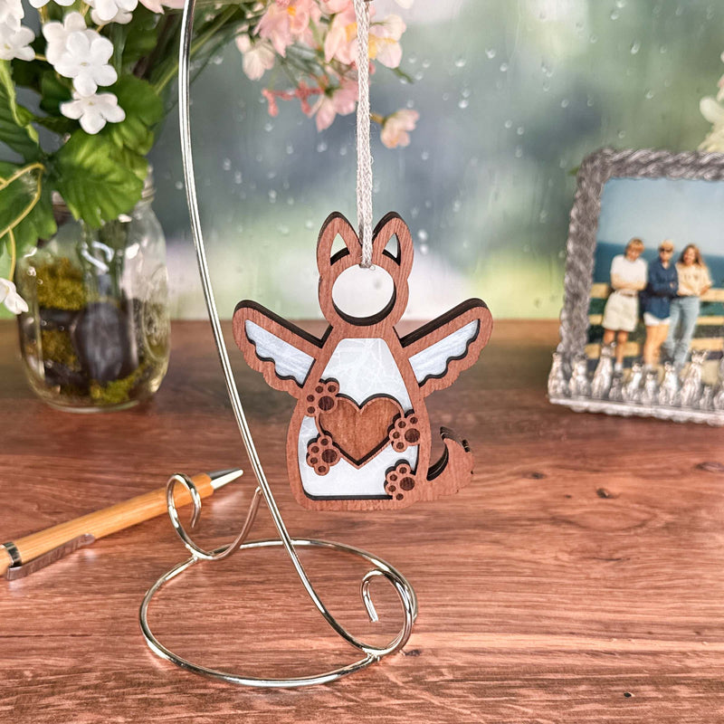 The Forged Flare® Forever By My Side Dog Angel Ornament, shaped like a winged dog holding a heart, hangs from a metal stand on a wooden table—an ideal dog lover gift or touching dog memorial.