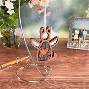 The Forever By My Side Dog Angel Ornament by Forged Flare® is a wooden winged dog with a heart, hanging on a silver stand—a thoughtful dog memorial or personalized keepsake, shown with flowers, a pen, and a beach photo in the background.