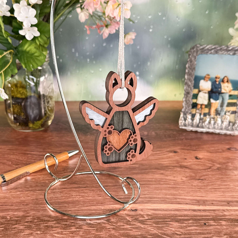 The Forever By My Side Dog Angel Ornament by Forged Flare® is a wooden winged dog with a heart, hanging on a silver stand—a thoughtful dog memorial or personalized keepsake, shown with flowers, a pen, and a beach photo in the background.