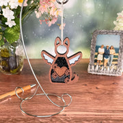 The Forged Flare® Forever By My Side Dog Angel Ornament, with cut-out heart and paw print wings, sits on a silver stand—an ideal dog lover gift or memorial. Shown on a wooden table with flowers, a framed photo, and pen in the background.