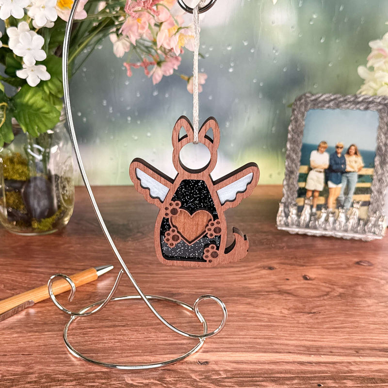 The Forged Flare® Forever By My Side Dog Angel Ornament, with cut-out heart and paw print wings, sits on a silver stand—an ideal dog lover gift or memorial. Shown on a wooden table with flowers, a framed photo, and pen in the background.