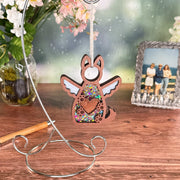 The Forever By My Side Dog Angel Ornament by Forged Flare® features a glittery, winged dog shape hanging on a metal stand—an ideal dog lover gift or memorial. A vase of flowers and a framed outdoor photo appear in the background.
