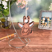 The Forged Flare® Forever By My Side Dog Angel Ornament, shaped like a wooden angel with wings and paw prints, hangs on a metal stand beside a pen, vase of flowers, and framed photo. A softly blurred background brings warmth.