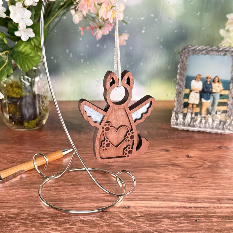 The Forged Flare® Forever By My Side Dog Angel Ornament, shaped like a wooden angel with wings and paw prints, hangs on a metal stand beside a pen, vase of flowers, and framed photo. A softly blurred background brings warmth.