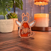 The Forged Flare® Forever By My Side Dog Angel Ornament stands on a wooden surface among potted plants and a lit candle—a cozy, warmly lit display ideal as a thoughtful dog lover gift or personalized decor.