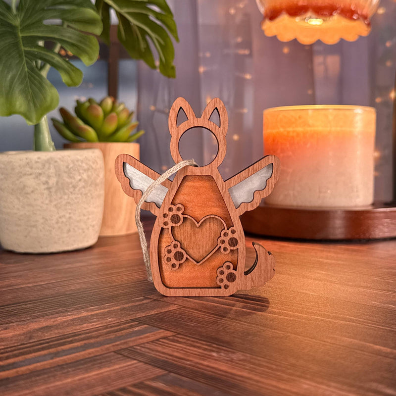 The Forever By My Side Dog Angel Ornament by Forged Flare®—a wooden figurine with wings, paw prints, and a heart cutout—stands on a wood surface, surrounded by a potted plant, succulent, candle, and glowing string lights.