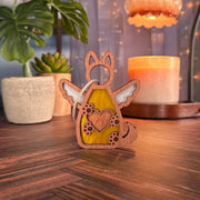 The Forged Flare® Forever By My Side Dog Angel Ornament, a wooden piece featuring wings, a heart cutout, and paw prints, sits on a wooden surface with candles and plants—perfect as a dog lover gift or personalized dog ornament.