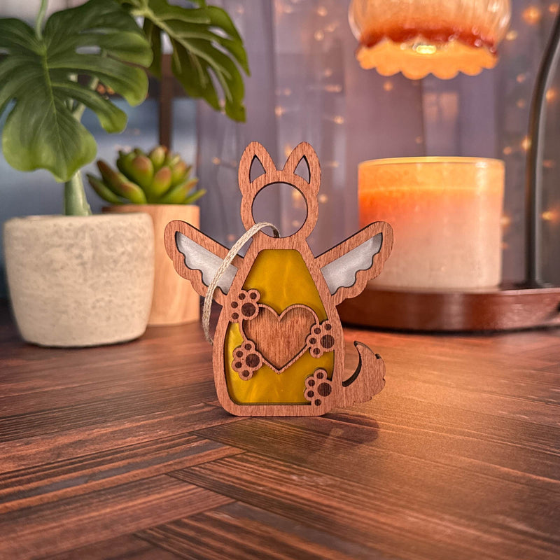 The Forged Flare® Forever By My Side Dog Angel Ornament, a wooden piece featuring wings, a heart cutout, and paw prints, sits on a wooden surface with candles and plants—perfect as a dog lover gift or personalized dog ornament.