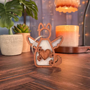 The Forged Flare® Forever By My Side Dog Angel Ornament, featuring wings and a heart, rests on a wooden surface. Potted plants, a lit candle, and string lights in the background create a cozy dog lover gift setting.