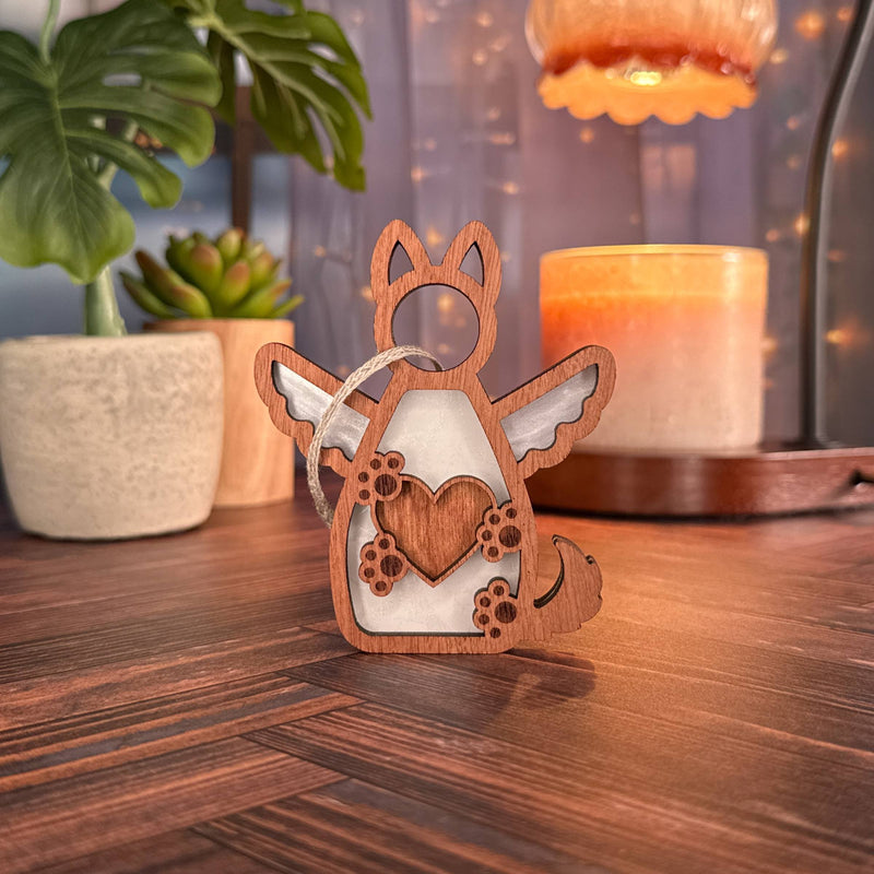 The Forged Flare® Forever By My Side Dog Angel Ornament, featuring wings and a heart, rests on a wooden surface. Potted plants, a lit candle, and string lights in the background create a cozy dog lover gift setting.