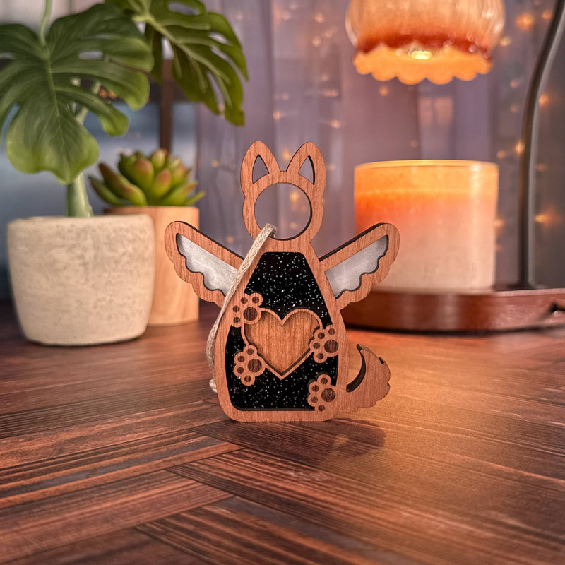The Forged Flare® Forever By My Side Dog Angel Ornament stands on a wooden surface with potted plants, a glowing candle, and string lights—creating a cozy scene great for pairing with other dog lover gifts or personalized ornaments.