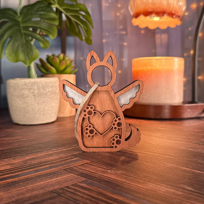 The Forged Flare® Forever By My Side Dog Angel Ornament—a wooden dog-shaped figurine with angel wings and a heart with paw prints—sits on a table, surrounded by cozy decor; it's the perfect dog lover gift or personalized ornament.