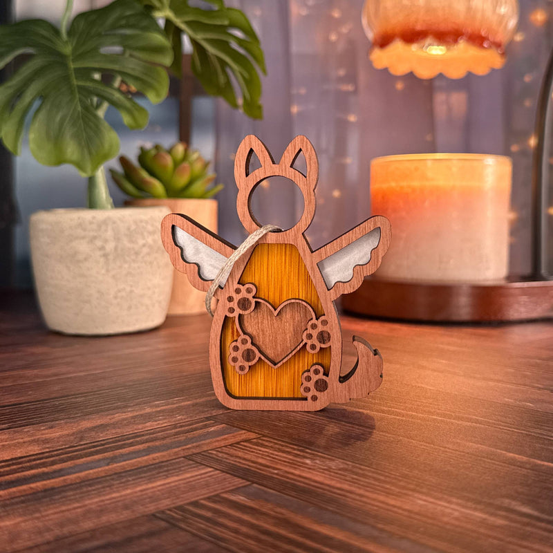 The Forged Flare® Forever By My Side Dog Angel Ornament—a small wooden figurine with wings and a heart—sits on wood, surrounded by potted plants and warm lighting. Perfect as a personalized dog ornament or meaningful gift for dog lovers.