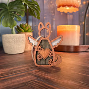 The Forged Flare® Forever By My Side Dog Angel Ornament, with wings and a heart, sits on a wooden surface—an ideal gift for dog lovers. Potted plants, a lit candle, string lights, and a glowing lamp add cozy ambiance in the background.